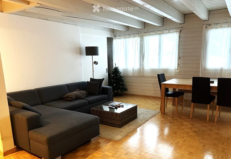 Bright living room with wooden flooring, large windows, grey sectional sofa, coffee table, dining table with chairs, floor lamp, and a small Christmas tree.