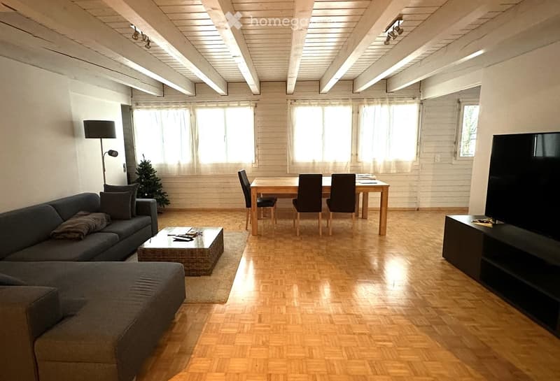 Bright living room with wooden flooring, exposed wooden ceiling beams, large windows with white curtains, a grey sectional sofa, a wicker coffee table, a dining table with chairs, a flat-screen TV on a stand, and a small Christmas tree in the corner.
