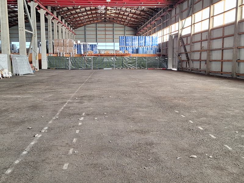 Empty warehouse space with concrete floor, metal walls, and scattered storage items including barrels and crates.