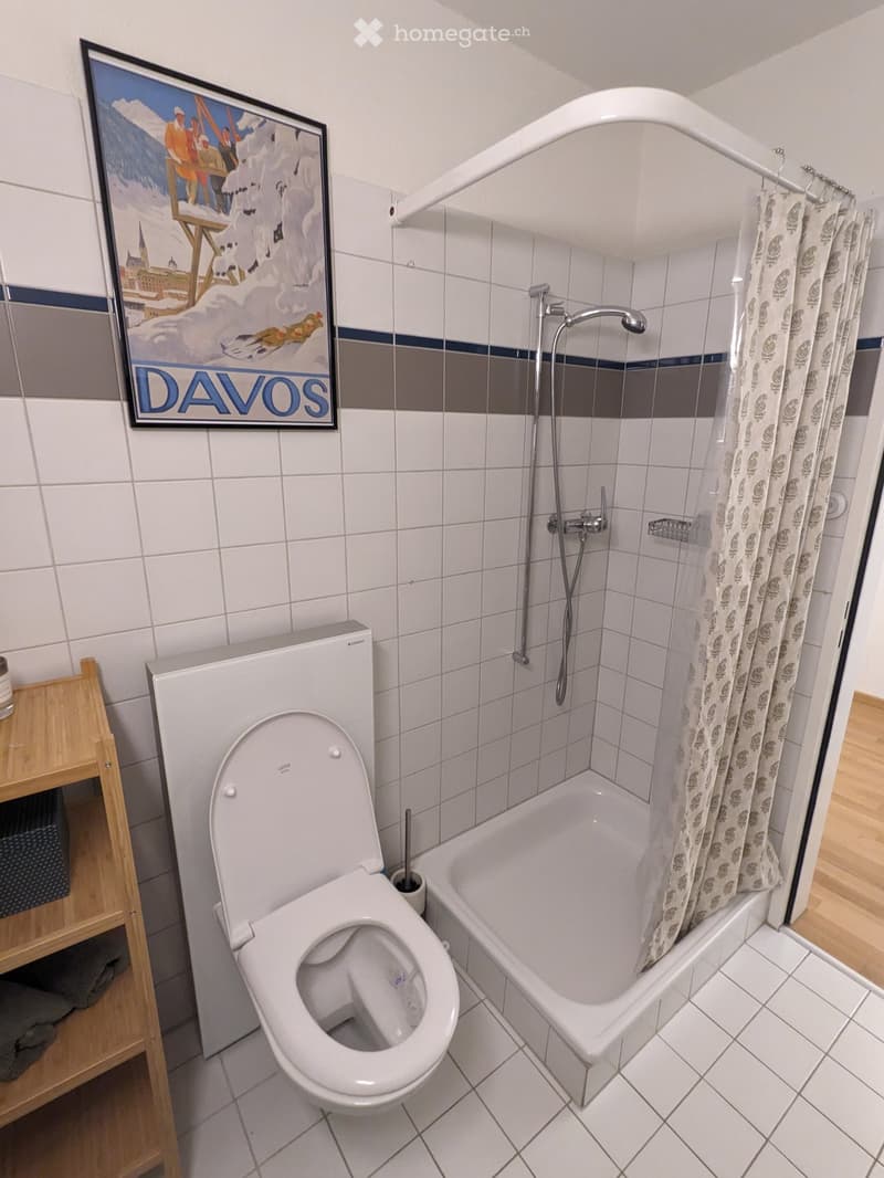 Bathroom with white tiles, a toilet, a bathtub with a shower curtain, a showerhead, and a wooden shelf with a basket.