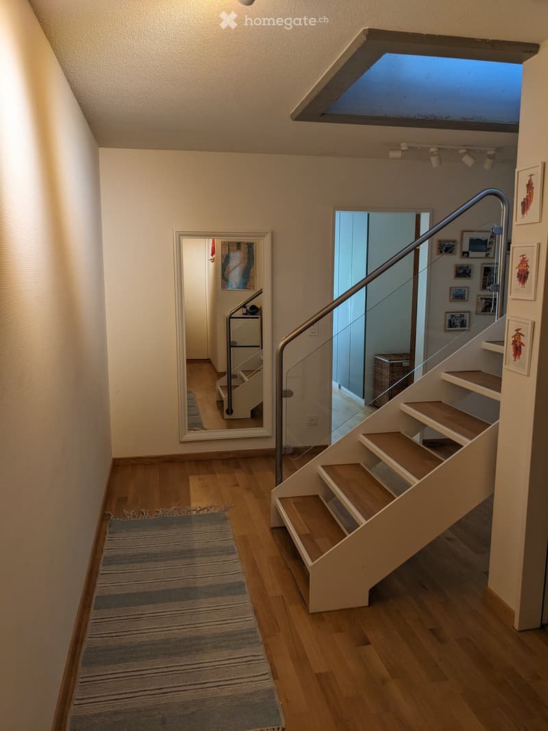 Modern hallway with wooden flooring, a staircase with glass railing, and a large mirror on the wall. The hallway features a striped rug and several framed pictures on the walls.