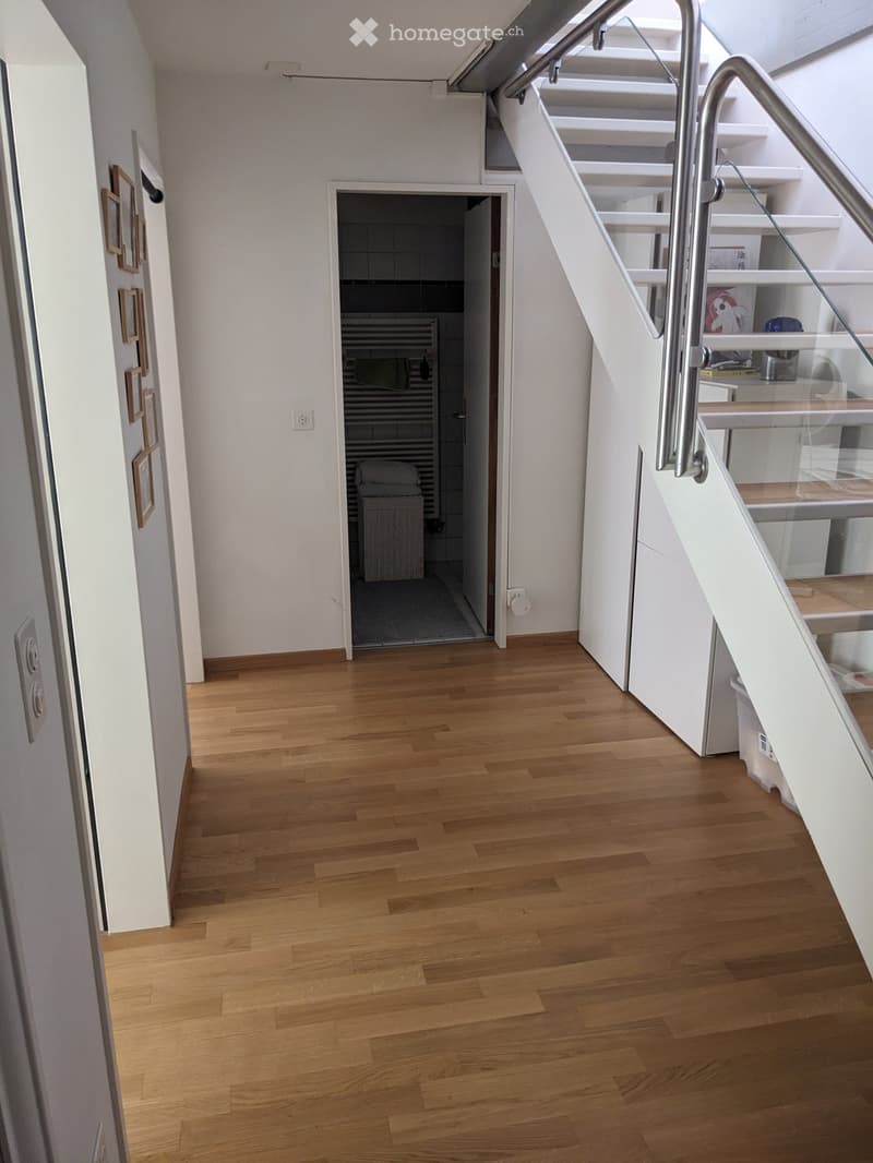 Bright hallway with wooden flooring, white walls, and a staircase with a glass railing leading to an upper level. A doorway leads to a tiled bathroom area with a visible radiator and storage basket.