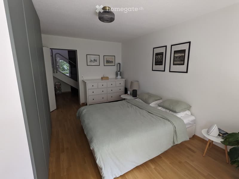 A modern, minimalist bedroom with a large bed, nightstand, lamp, drawer unit, and framed pictures on the wall. The room features light wooden flooring and a small table with a plant in the corner.