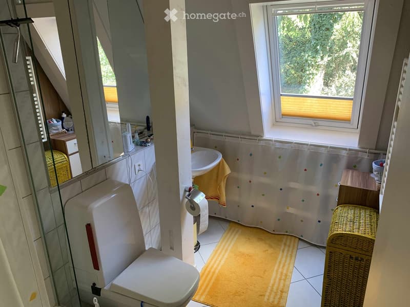 The bathroom features a toilet, sink, mirror, shower curtain, and a wicker basket. The space is well-lit with natural light from a window and has a yellow bath mat on the floor.