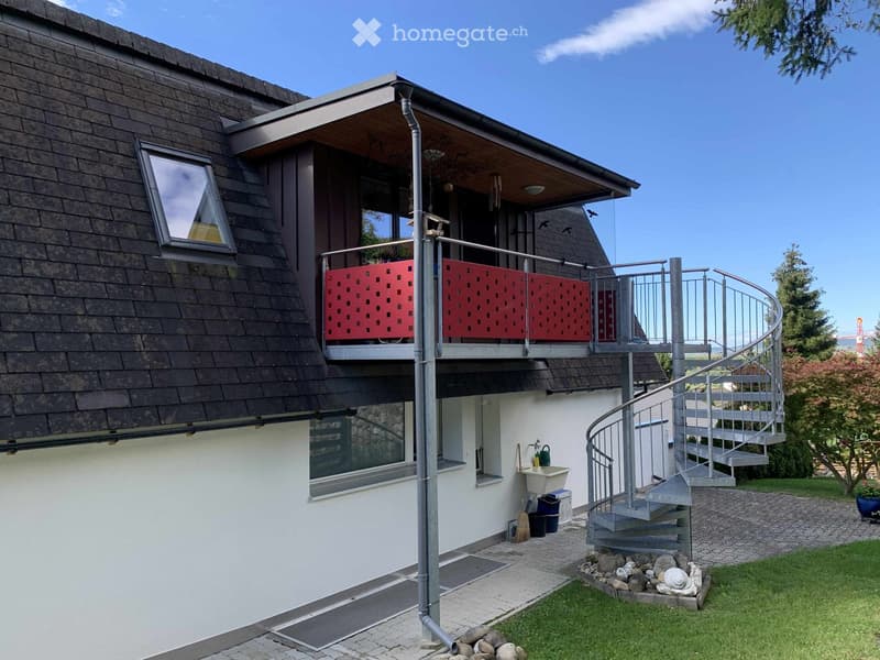 Two-story house with a modern design, featuring a balcony with red railings, a spiral staircase leading to the garden, and a small outdoor sink area.