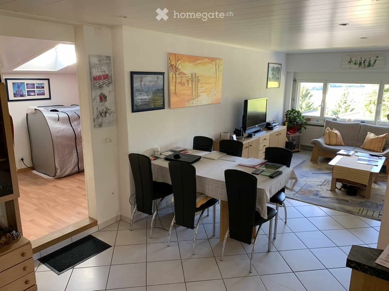 Open-plan living and dining area with a sofa, coffee table, dining table with chairs, laptop, TV, and a plant. The space features a mix of tiled and wooden flooring, large windows, and artwork on the walls.