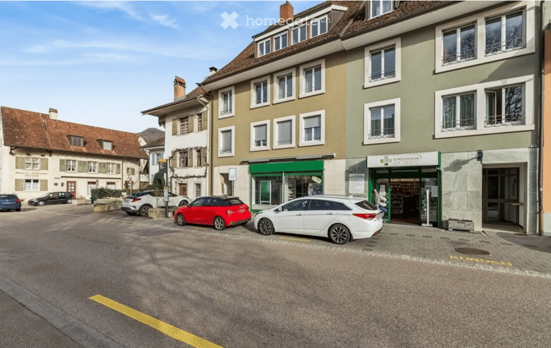 Multi-story building with multiple windows and a storefront on the ground floor, surrounded by parked cars and a paved street.