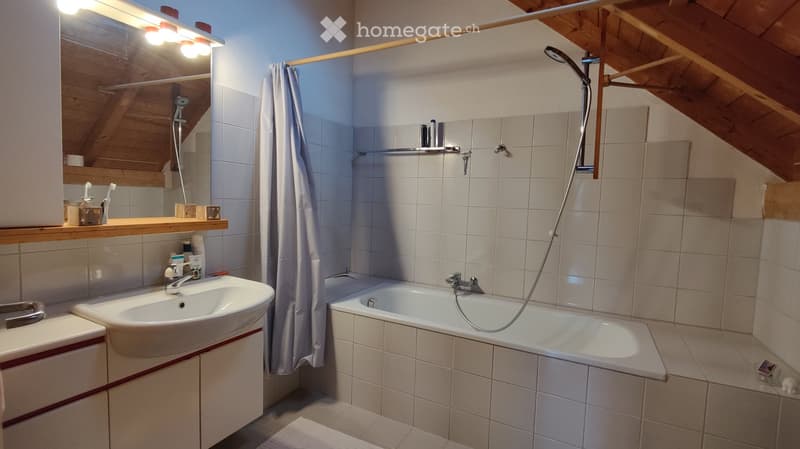 Cozy bathroom with a sink, bathtub, and shower area. Features a wooden shelf with toiletries, a mirror with built-in lighting, and a slanted wooden ceiling.