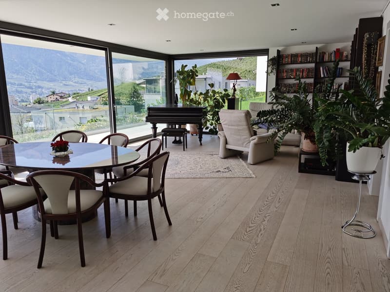 Spacious room with wooden flooring, large windows offering a scenic mountain view, round dining table with chairs, sofa, piano, bookshelf, and potted plants.