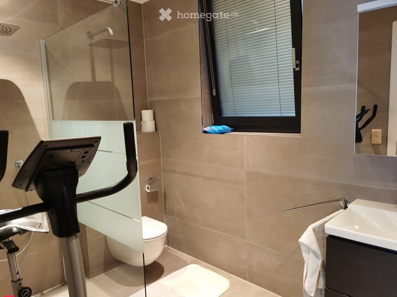 Modern bathroom with beige tiles, a toilet, a sink with a mirror, a towel, and an exercise bike in the corner.