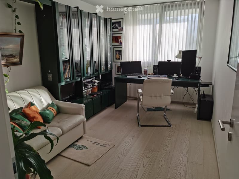 A modern, well-lit room with a white leather sofa, a desk with two computer screens, an office chair, a bookshelf, and a plant. The room features wooden flooring and large windows with sheer curtains.
