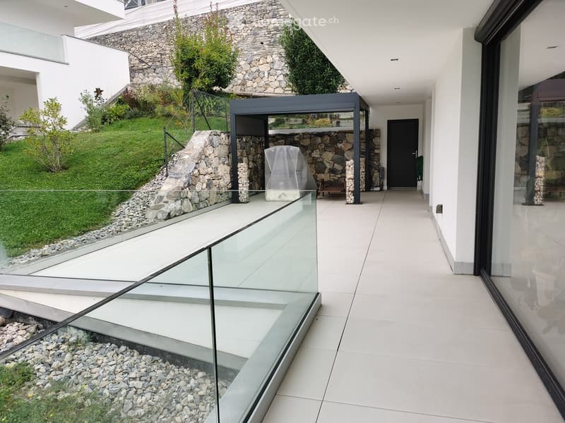 Modern hallway with white tiled flooring, glass railings, and a raised ground floor leading to a landscaped garden area with stone walls and greenery.