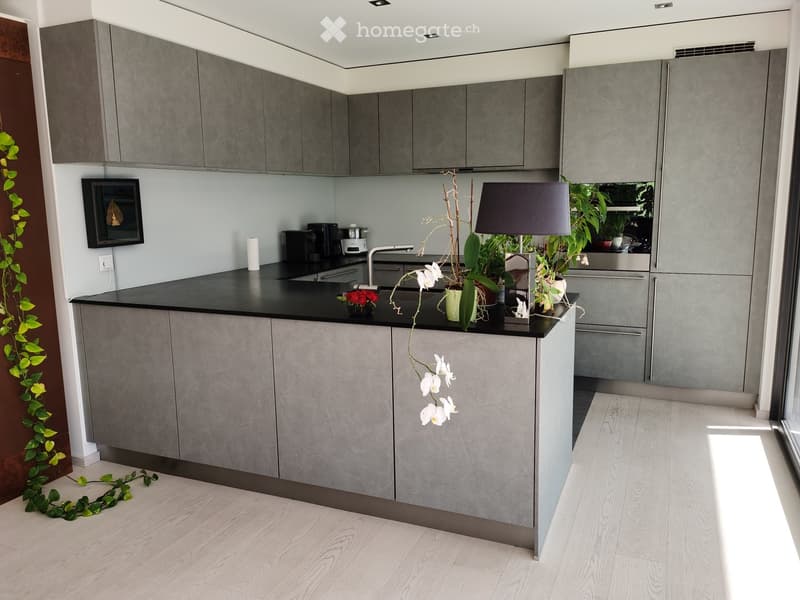 Modern kitchen with grey cabinets, black countertop, and a central island. Features a faucet, sink, coffee maker, oven, fridge, table lamp, and decorative flower vases.