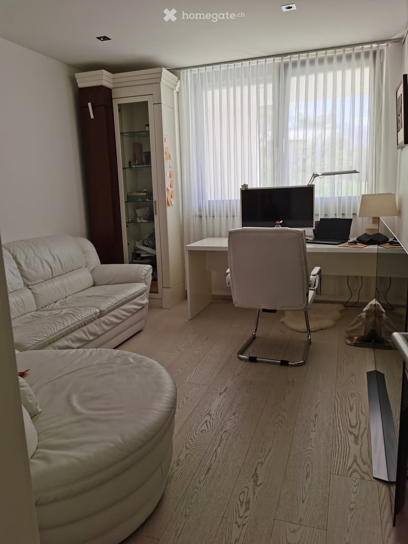 A modern, well-lit room with a sofa, ottoman, desk, chair, laptop, screen, lamp, and cabinet. The room features wooden flooring and large windows with sheer curtains.