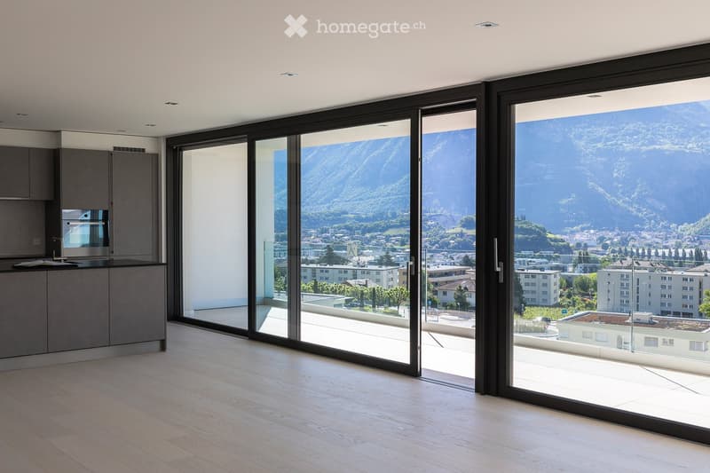 Modern kitchen with light wood flooring, grey cabinets, and expansive floor-to-ceiling windows offering a scenic mountain view.