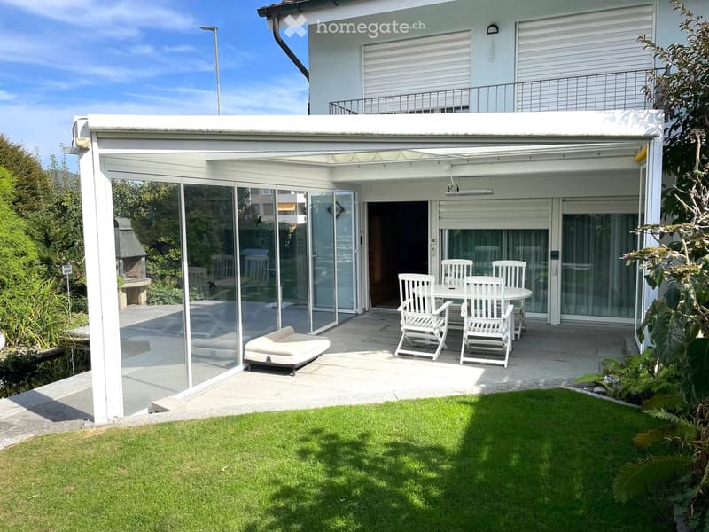 Garden with a covered patio area, white furniture including a table, chairs, and a sofa, surrounded by greenery and a well-maintained lawn.