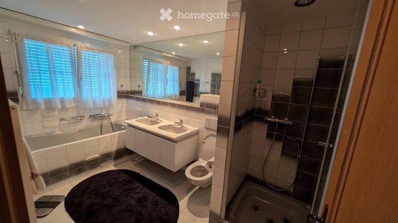Modern bathroom with a bathtub, double sink, toilet, shower, and large mirror. Features tiled walls, a dark round rug, and a window with blinds.