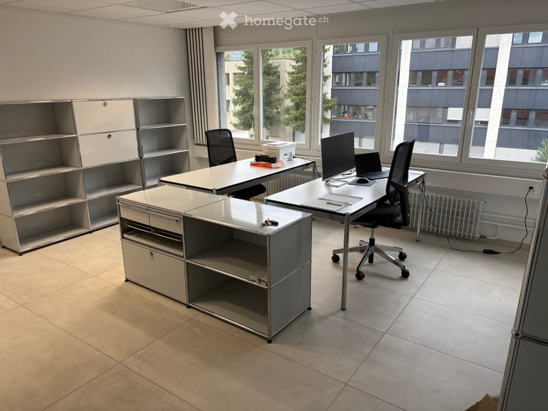 Modern office space with two desks, office chairs, laptops, a screen, and a cabinet. Large windows provide natural light, and the floor is tiled.