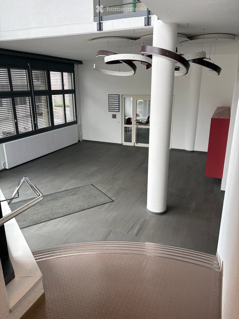 Modern hallway with grey tiled floor, white walls, large windows, and modern lighting fixtures. Features a staircase with a metal railing and a red accent wall.
