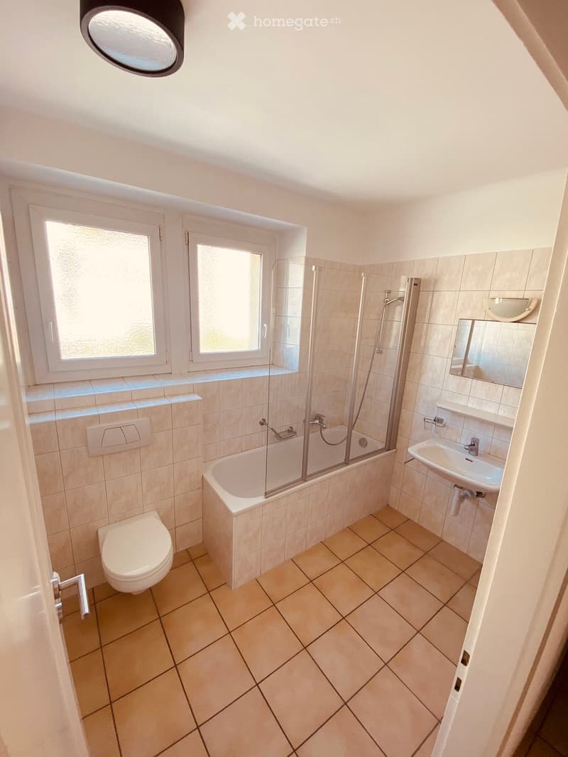 Modern bathroom with beige tiles, a bathtub with a glass shower enclosure, a toilet, a sink with a mirror, and a window providing natural light.