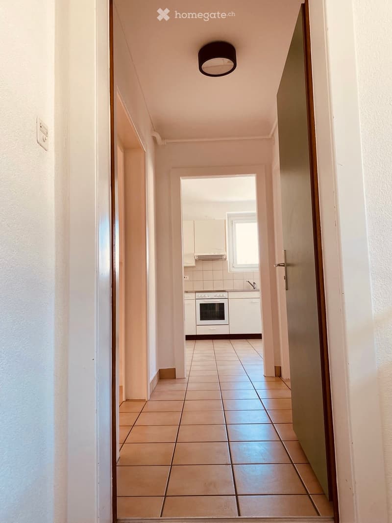 A narrow hallway with tiled flooring, white walls, and a ceiling light fixture. A door on the right leads to a kitchen area with white cabinets, a built-in oven, and a window.