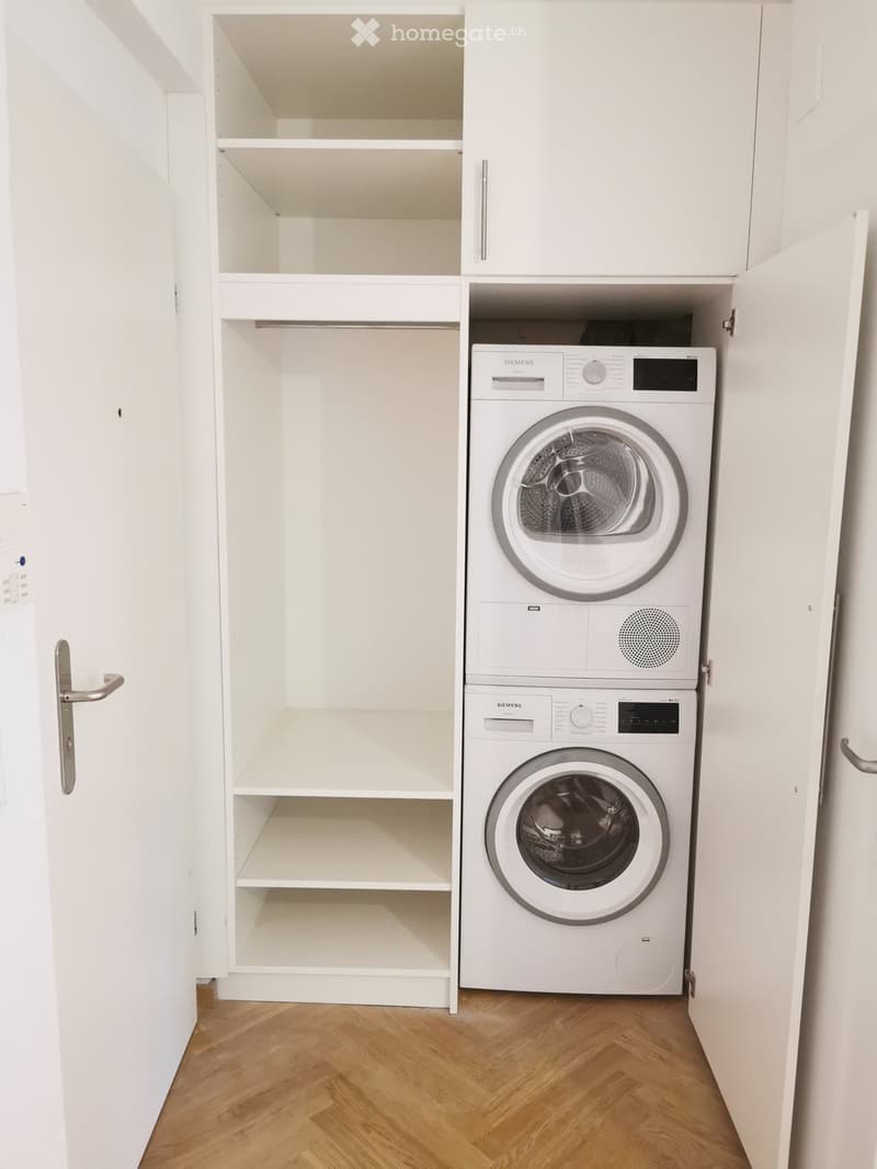 A small, modern laundry area with a white washing machine and dryer stacked vertically, white built-in shelving, and wooden flooring.