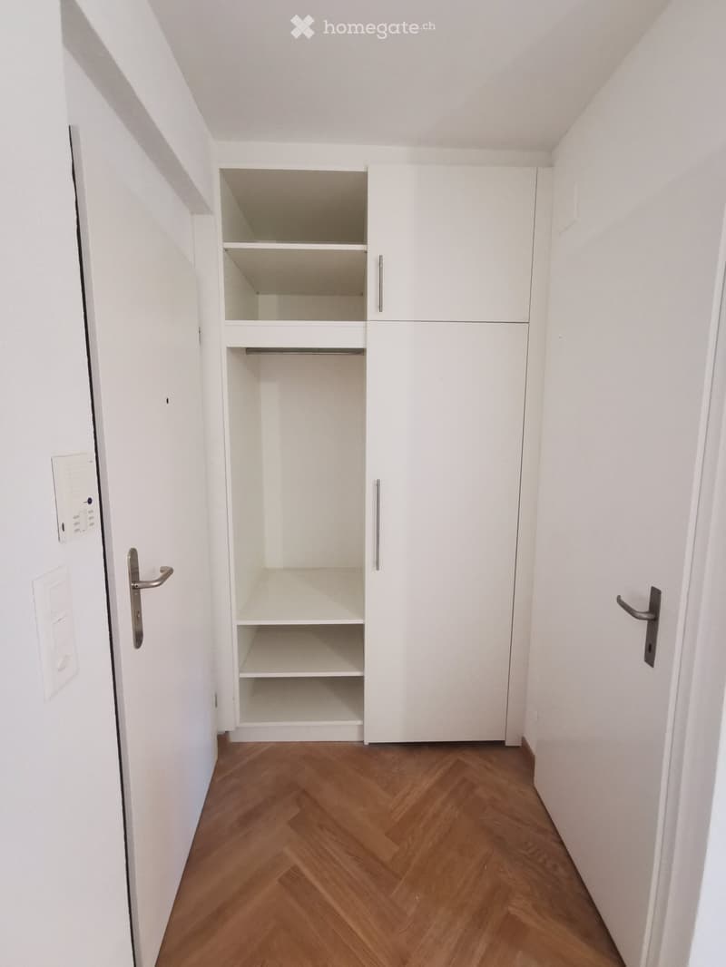 Empty hallway with white walls, wooden floor, and a built-in white closet with open shelves and drawers.
