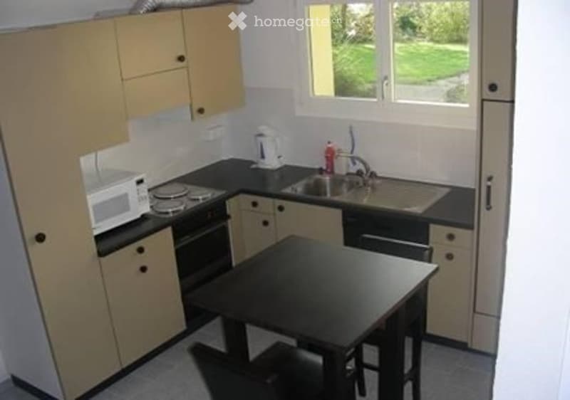 Compact kitchen with light wood cabinets, black countertop, microwave, oven, sink, and a small dining table with chairs.