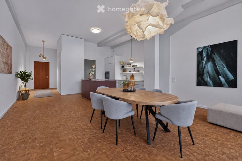 Modern dining area with a round wooden table surrounded by grey chairs, pendant lighting, and a small kitchen area with cabinets and a countertop.