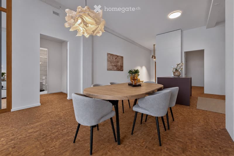 Modern dining room with a round wooden dining table surrounded by grey upholstered chairs, a decorative pendant light, and a small bar area with a vase and potted plants.