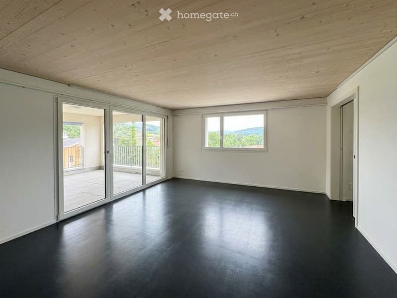 Empty room with white walls, dark flooring, large sliding glass doors leading to a balcony with a scenic view, and a window with a view of greenery and mountains.