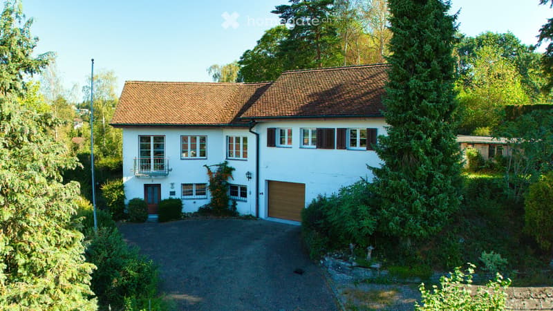 Two-story white house with a brown tiled roof, multiple windows, a garage, and a driveway surrounded by greenery.