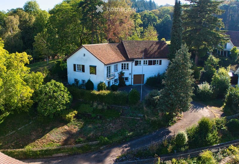 A two-story white house with a brown tiled roof, multiple windows, and a garage, surrounded by lush greenery and trees.