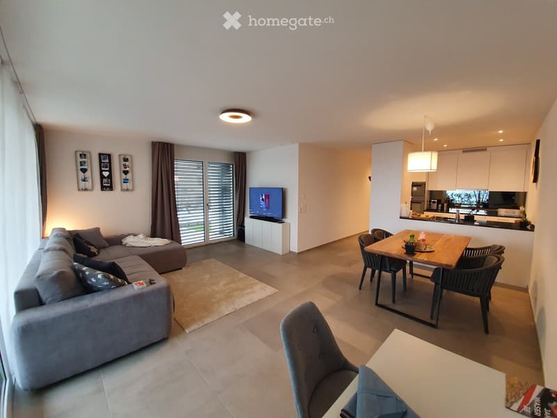 Modern open-plan living and dining area with a grey sectional sofa, coffee table, dining table with chairs, television, and a small kitchen area with cabinets and appliances.