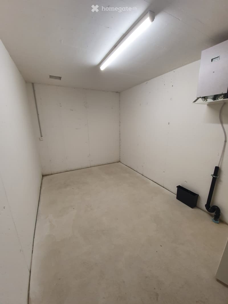 Empty room with bare walls and concrete floor, featuring a small window with a light fixture above it and a white appliance mounted on the wall.