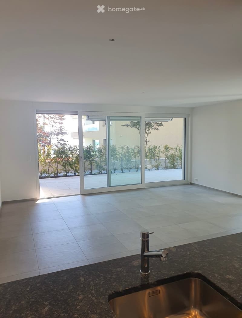 Modern kitchen with granite countertop, stainless steel sink, and faucet. Large sliding glass doors open to a tiled patio with greenery.