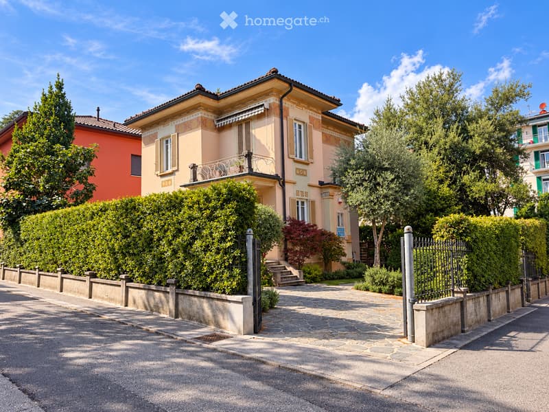 Two-story house with beige and pink facade, decorative shutters, and a small balcony. The house is surrounded by greenery and a stone pathway leads to the entrance.