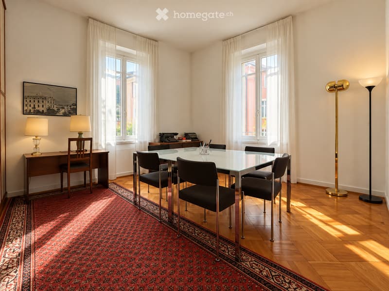 Brightly lit room with wooden flooring, large windows with white curtains, a glass dining table surrounded by chairs, a wooden desk with a lamp, and two tall floor lamps.