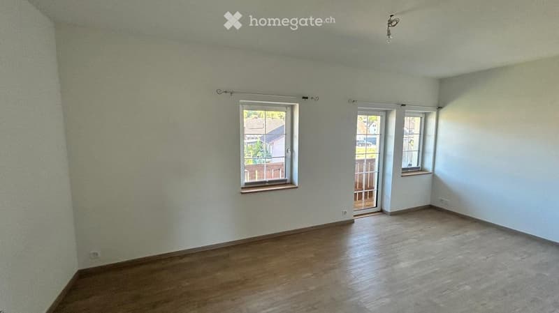 Empty room with light-colored walls, wooden flooring, and large windows, including a sliding glass door leading outside.