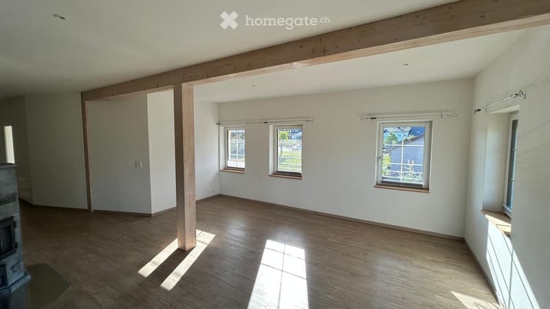 Empty room with wooden flooring, white walls, and multiple windows allowing natural light to enter. The room features exposed wooden beams on the ceiling and a small radiator on the left side.
