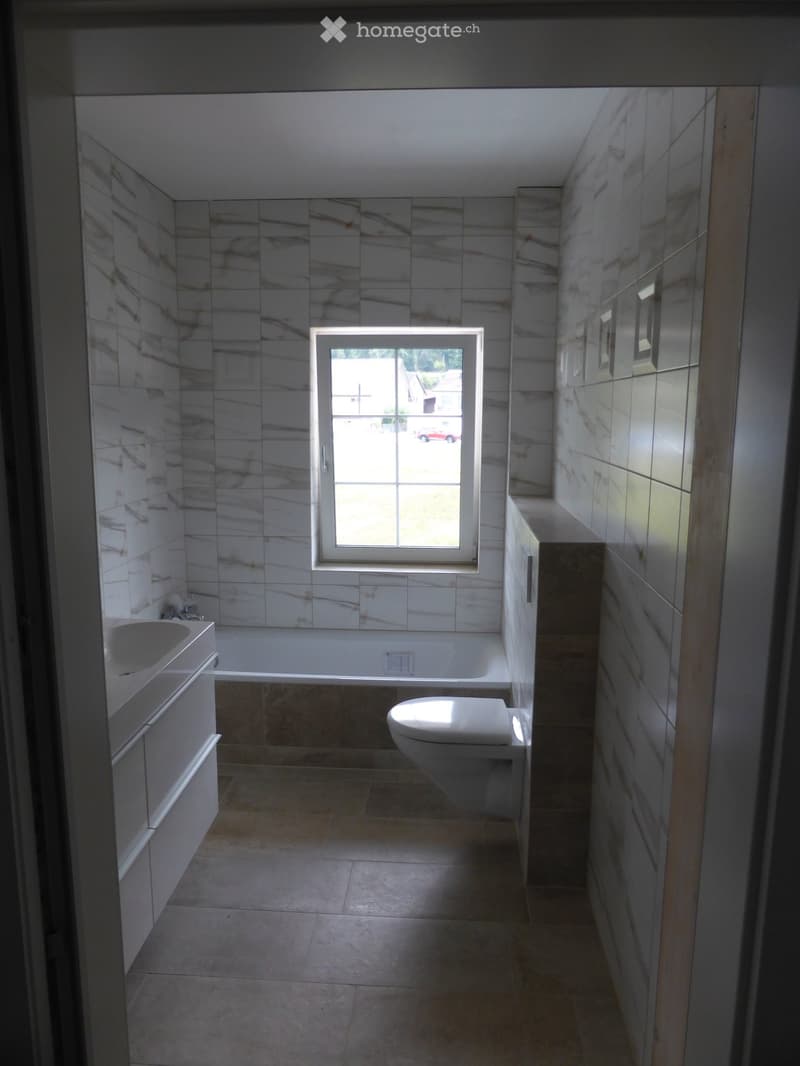 Modern bathroom with marble tiles, a bathtub, toilet, sink, and a window providing natural light.