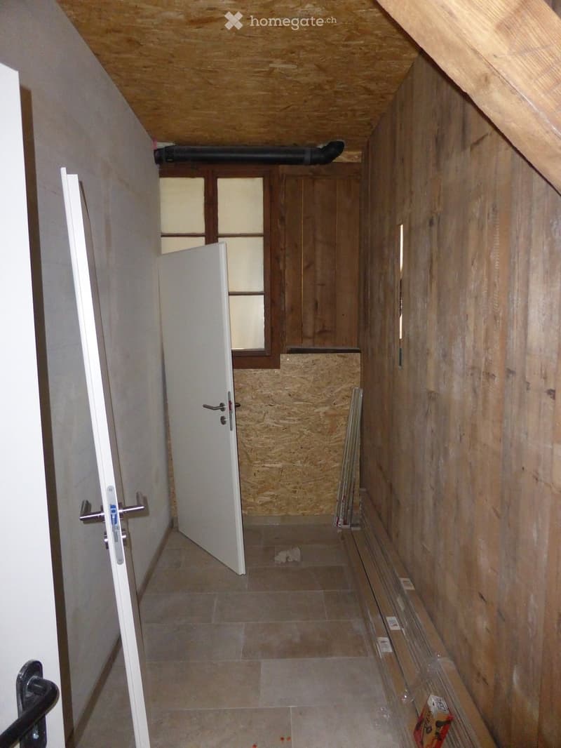 Empty hallway with wooden walls, tiled floor, and a partially open door leading to another room. Exposed wooden beams on the ceiling and a window with frosted glass panes are visible.
