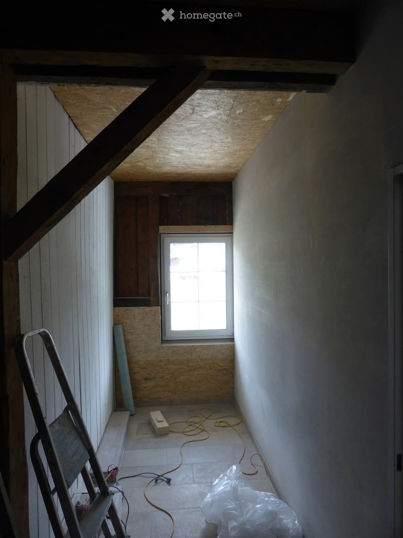 Empty room with exposed wooden beams, unfinished walls, and a window at the far end. Ladder and construction materials are present.