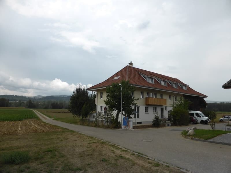 Two-story house with a red-tiled roof, white walls, and multiple windows. Features a balcony on the upper floor and a driveway with parked vehicles.