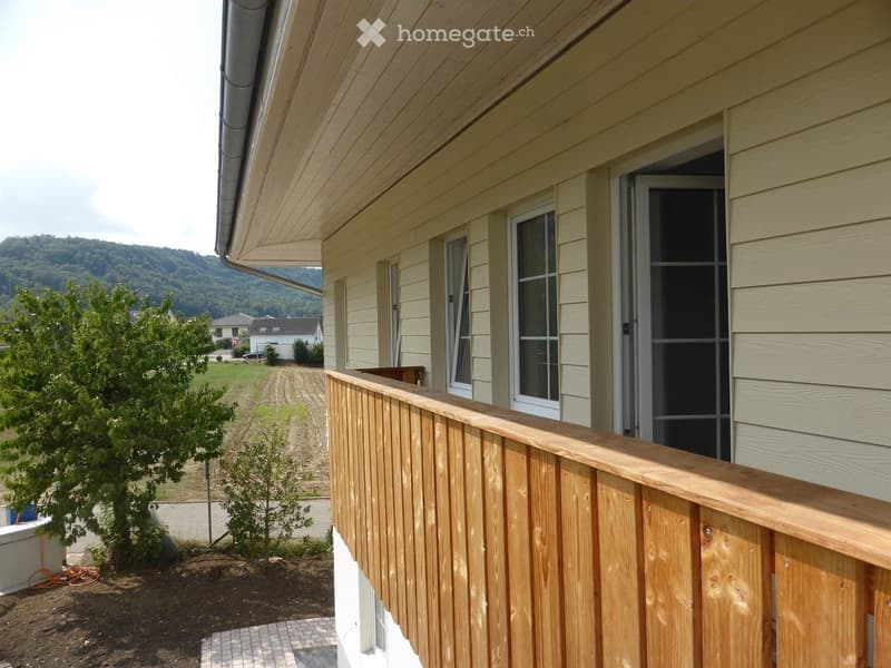 Wooden balcony railing with a view of a grassy yard, trees, and distant hills.