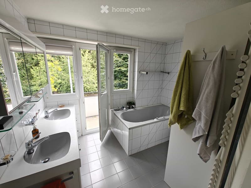 Modern bathroom with white tiles, double sink, bathtub, large windows offering a view of greenery outside, and towels hanging on a rack.