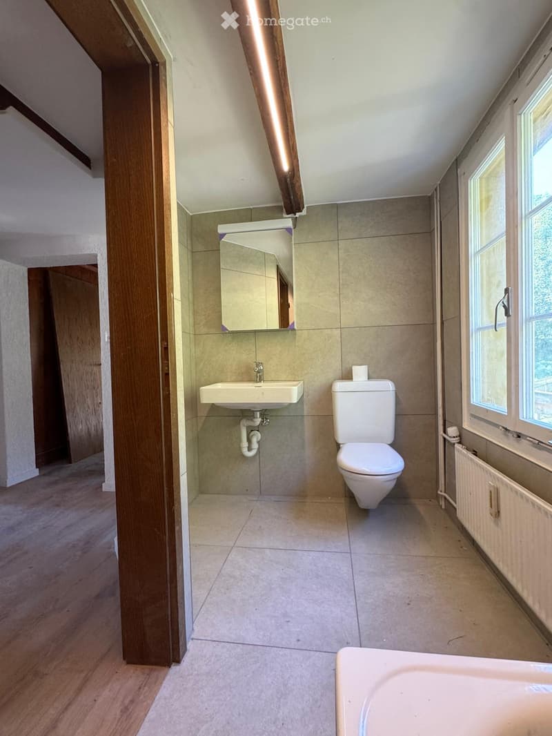 Modern bathroom with beige tiles, a toilet, a sink, and a mirror. The room features wooden flooring and a window providing natural light.