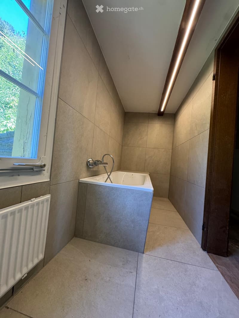 Modern bathroom with a corner bathtub, sleek faucet, and built-in radiator. The space features light-colored tiles and a large window providing natural light.