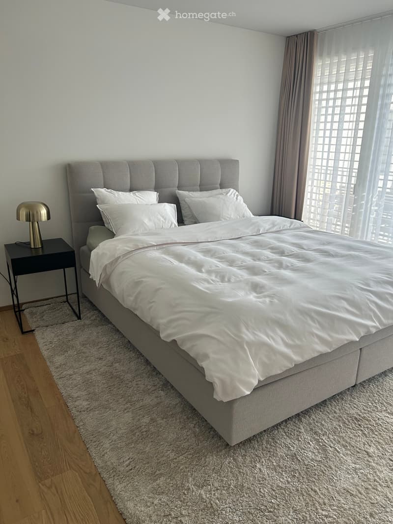 A modern bedroom with a large bed, white bedding, a tufted headboard, a nightstand with a lamp, and a shaggy rug on a wooden floor.
