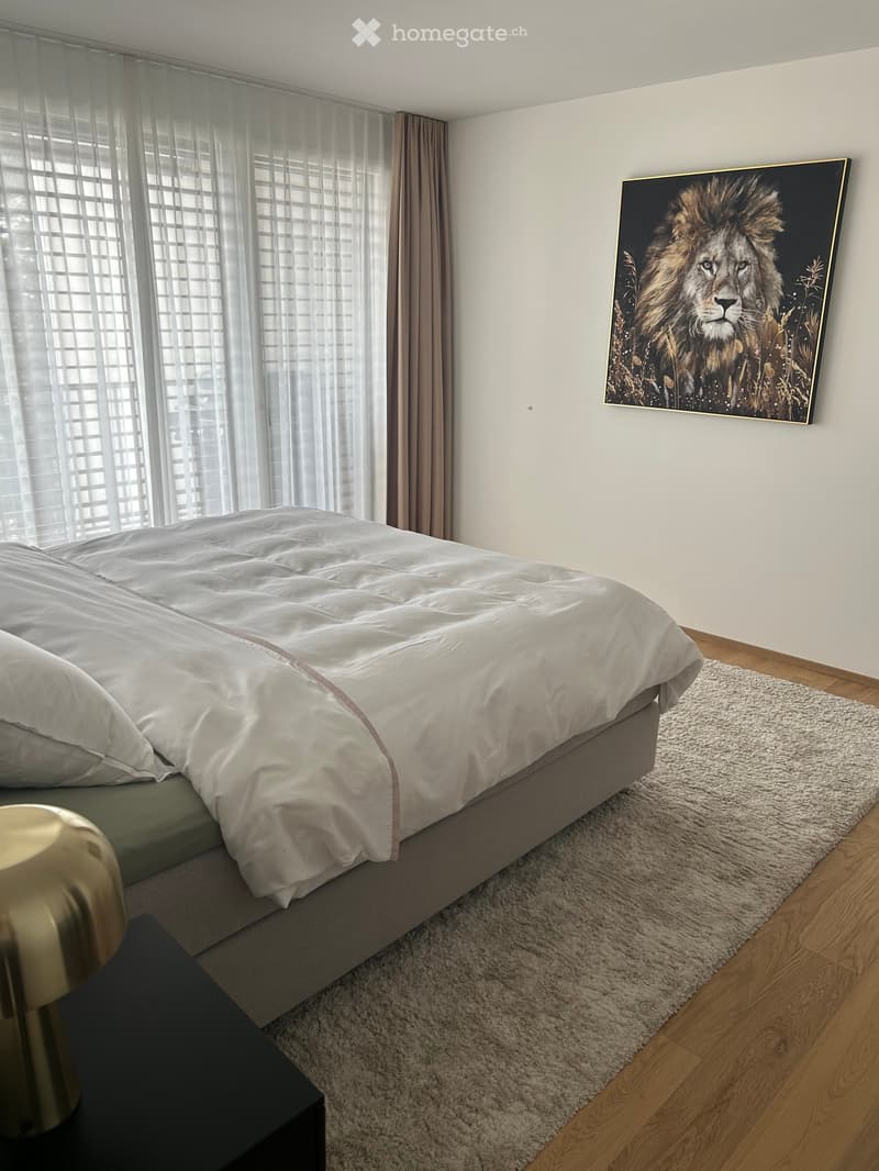 Modern bedroom with a large bed, nightstand, lamp, and a large painting of a lion on the wall. The room features wooden flooring and a white shaggy rug.